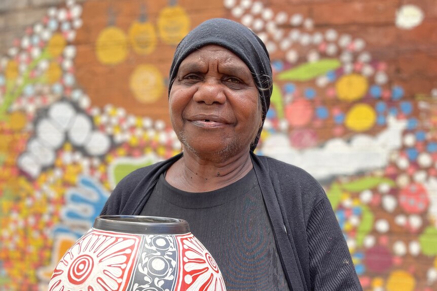An Aboriginal woman holds a beautifully decorated pot with Indigenous artwork behind her.