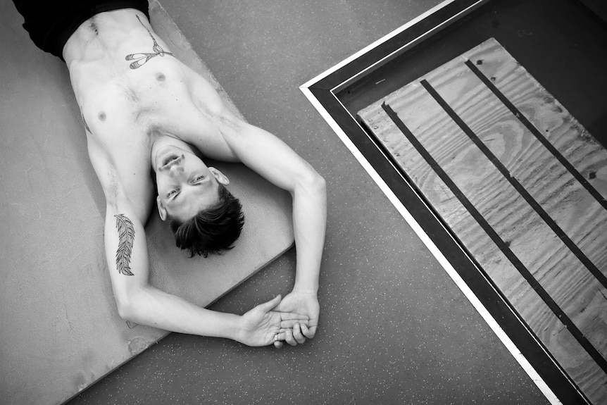 A young man lies on the floor breathing heavily after an exercise, with tattoos visible on his bare chest and arms. 