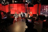 Four journalists sit on a red TV set surround by four large studio cameras.
