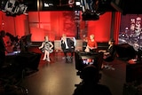 Four journalists sit on a red TV set surround by four large studio cameras.