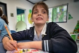 A picture of a young girl in a school uniform drawing on a piece of paper. She sits at a table in a classroom.