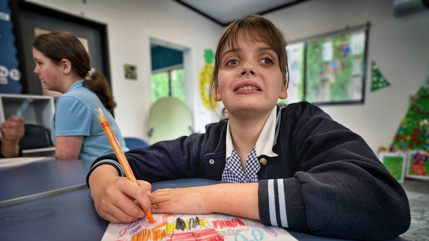 A picture of a young girl in a school uniform drawing on a piece of paper. She sits at a table in a classroom.