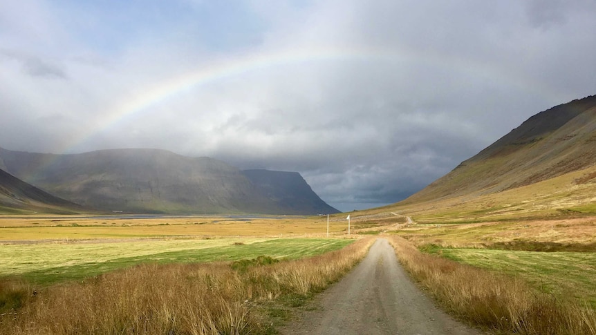 A faint rainbow stretches between two mountains, on top of an empty dirt road.