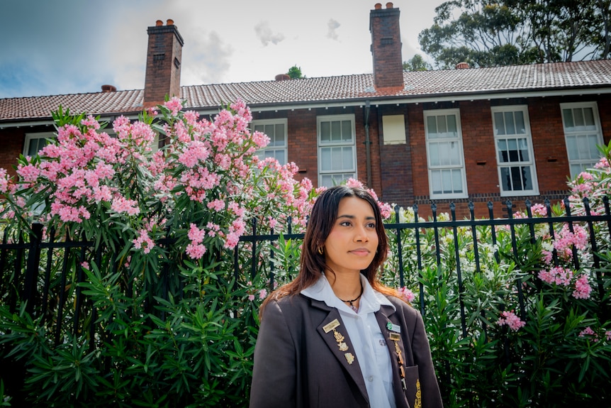 A girl with dark hair wearing a blue school uniform stands in front of a fence covered in bright pink flowers and red brick buil