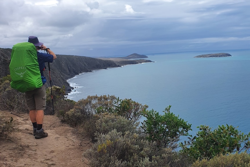 A man in hiking gear including a large backpack looks out to the ocean, standing on a cliff