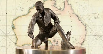 Brass Matthew Flinders statue on podium bent over with cat Trim against background map of Australia.