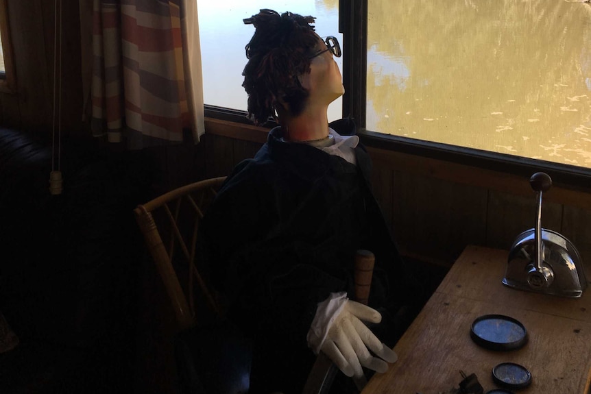 An effigy sits at a desk inside a houseboat looking out the window to the river.