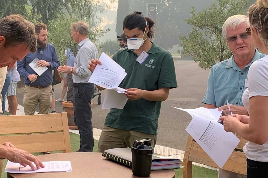 People gather under a smoky sky at a winery and sign paper work. One person wears a smoke mask.