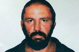 A mugshot-style photo of a man with dark hair, eyes and beard. He looks menacingly into the camera.