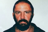 A mugshot-style photo of a man with dark hair, eyes and beard. He looks menacingly into the camera.