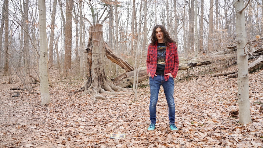 Kurt Vile in a forest during autumn, standing on fallen leaves. He wears a flannel shirt and jeans and has hands in pockets