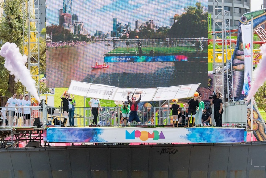 A woman with a primitive paraglider jumps off a stage into the water at an outdoor festival in front of a large TV screen.