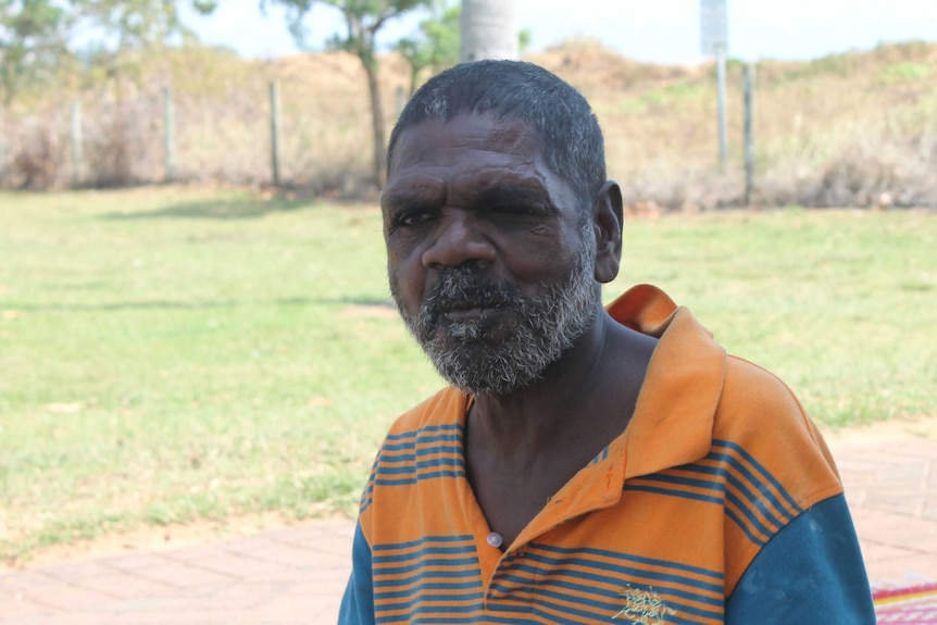 David Gurruwiwi squints with one eye, and wears and orange shirt as he sits on the grass