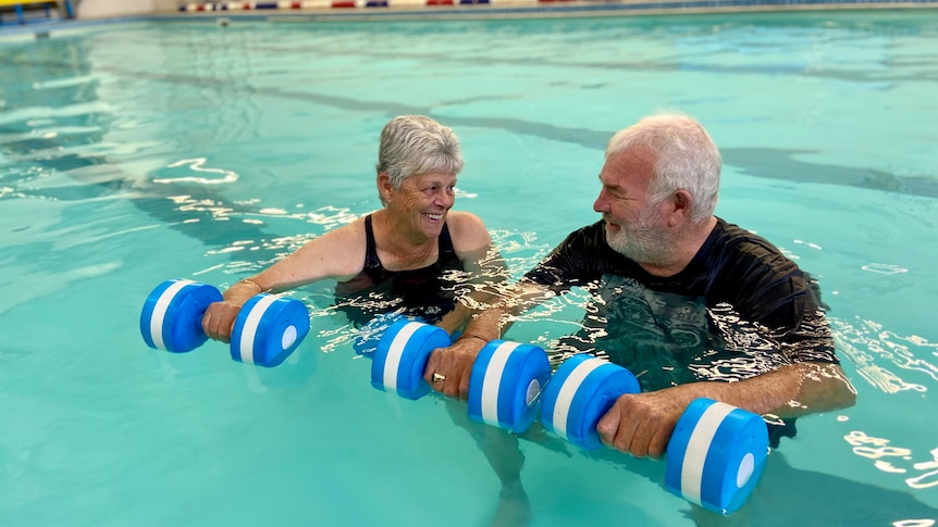 An elderly couple floating in a pool holding blue and white foam dumbbells' smile lovingly at each other.