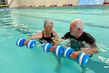 An elderly couple floating in a pool holding blue and white foam dumbbells' smile lovingly at each other.