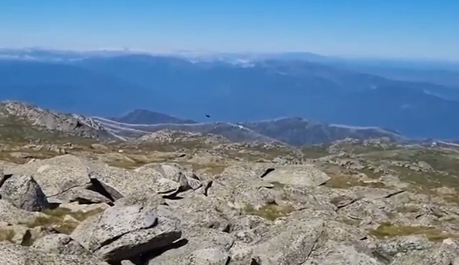 A scenic view of rocks and mountains with the silhouette of a fighter jet flying low in the bright blue sky.