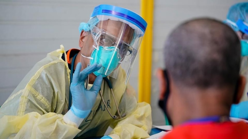 A man in PPE talks to a man in a red t-shirt at a desk