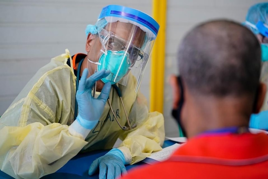 A man in PPE talks to a man in a red t-shirt at a desk