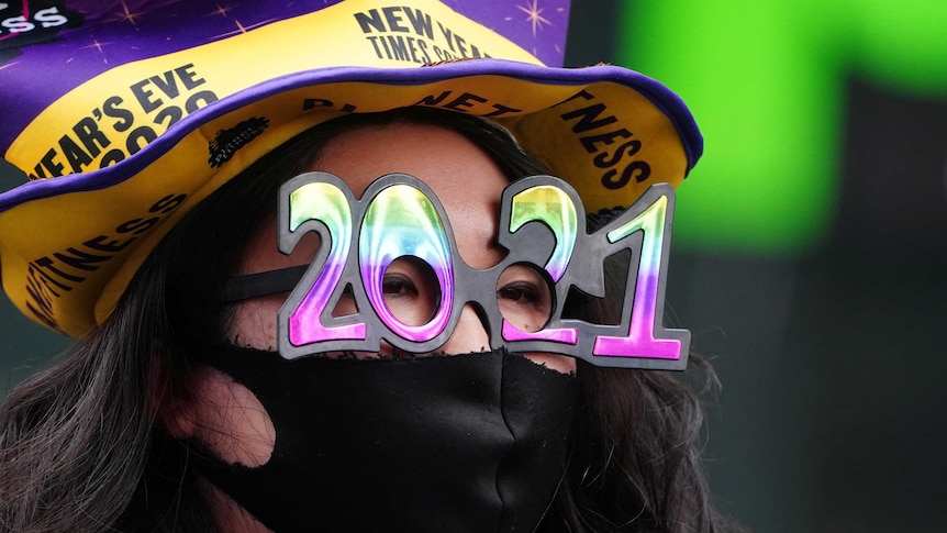 A woman wearing 2021 a face mask, glasses and a top hat with New Year's Eve 2020 written on it stares off camera