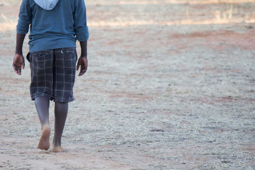 An Indigenous boy, whose face cannot be seen, walks barefoot in an outback area.