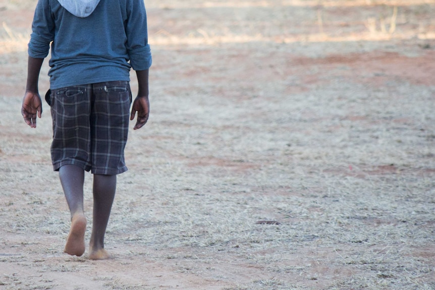 A young indigenous person shown from the shoulders down to feet walks on dirt road.