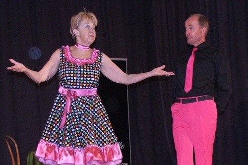 A blonde woman in pink performing on stage next to a man wearing pink trousers, black shirt and pink tie.
