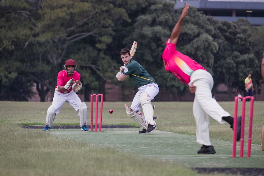 A batsman in a green shirt receives a ball from a bowler in a pink shirt.
