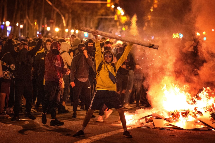 A protester stands with a large pipe held above his head as fire burns in the background