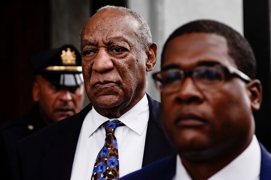 Bill Cosby wears a stony expression and a suit as he walks behind one man, and ahead of a uniformed police officer