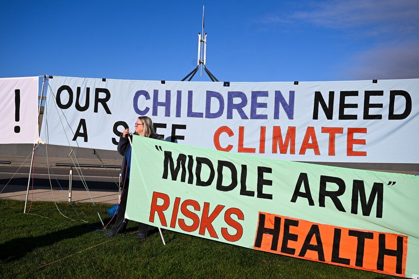 An activist holds a banner reading 'Middle Arm Risks Health' and 'Our Children Need A Safe Climate'