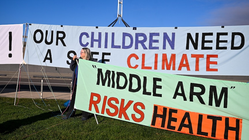 An activist holds a banner reading 'Middle Arm Risks Health' and 'Our Children Need A Safe Climate'