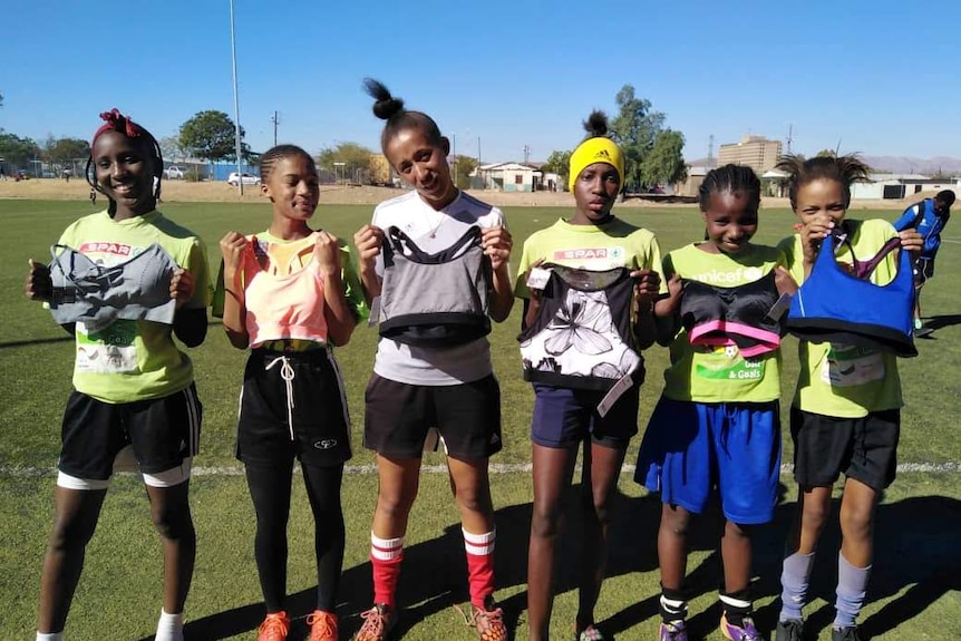 A group of young African women hold sports bras while standing on a soccer field.
