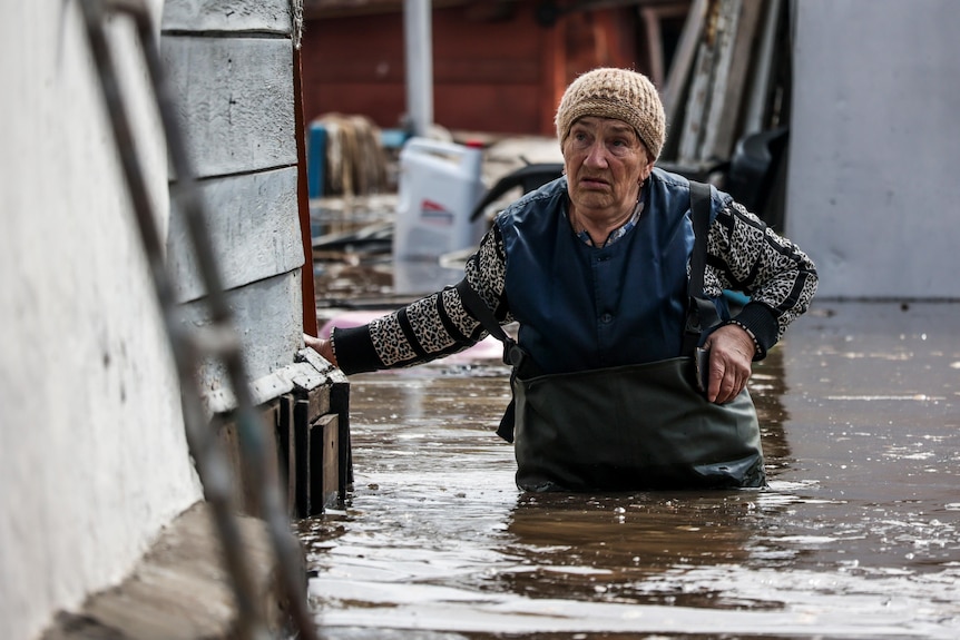 An elderly woman standing next to a house in flood waters up to her waist.