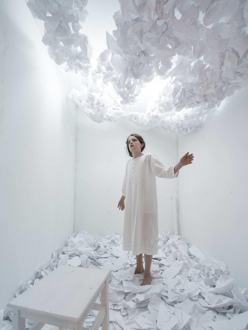 A girl in a white room with scrunched up paper on the ceiling and on the floor