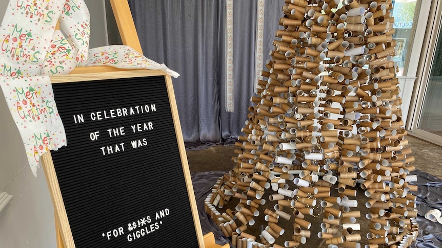 A black board with writing on it next to a christmas tree made entirely out of toilet paper rolls.