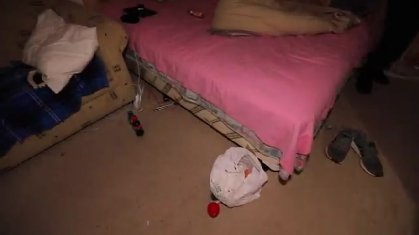 An unmade bed and couch with pillow and blanket, in a room with a plastic bag and shoes on floor.