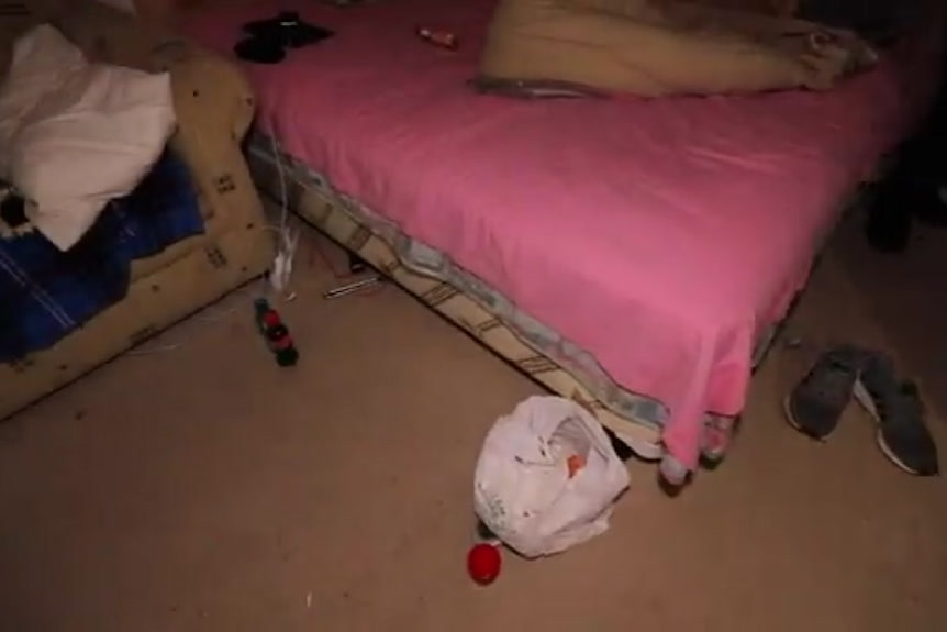 An unmade bed and couch with pillow and blanket, in a room with a plastic bag and shoes on floor.