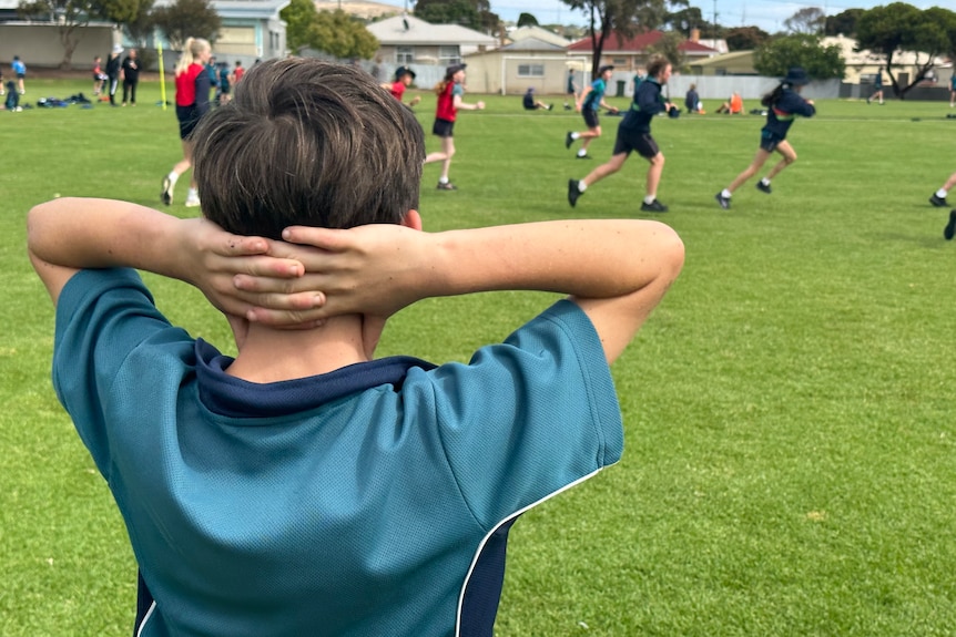 A small school age boy stressed with hands wrapped behind his head watching a game of football.