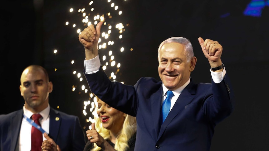 Benjamin Netanyahu wears a blue suit and has his thumbs up as fireworks go off in the background