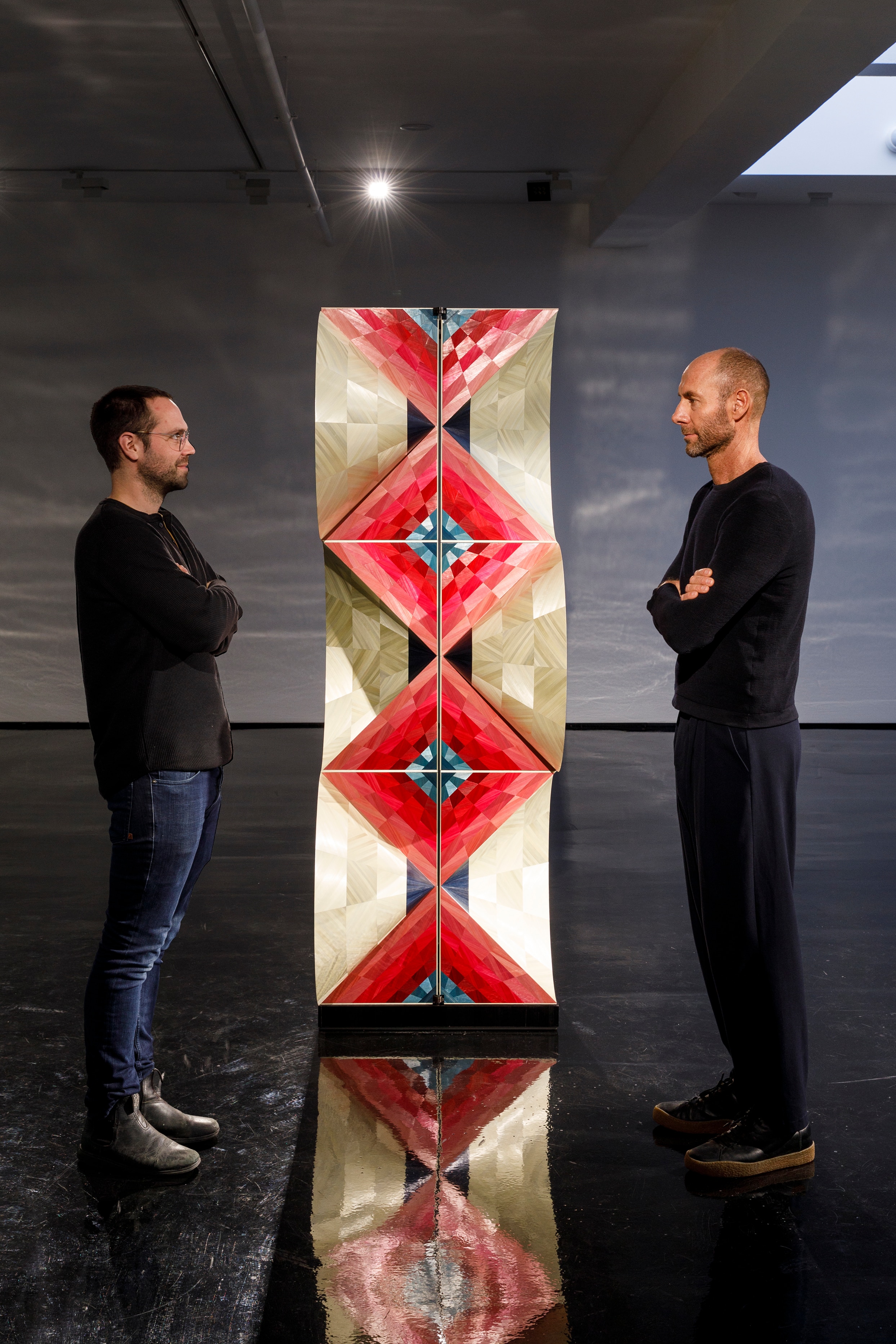 Two men stand on either side of an artwork - a thin box that stands tall, with an odd wavy illusion via its gold-red patterns