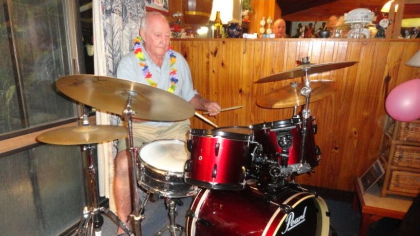 Colour photo of older man playing drums wearing a lei, timber wall behind and another man's head
