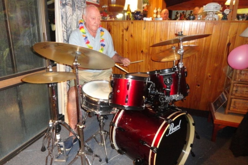 Colour photo of older man playing drums wearing a lei, timber wall behind and another man's head