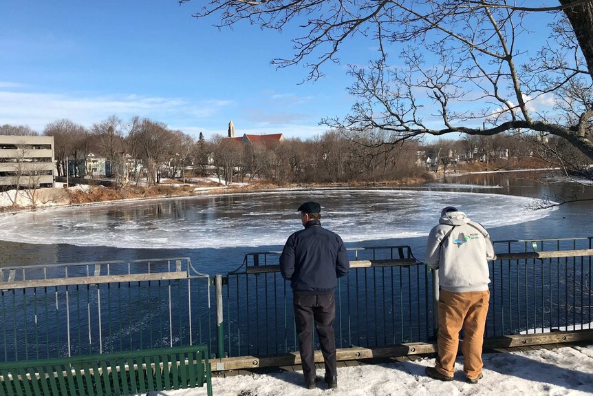 Two people stand on a river back watching a large disk of ice in a river with city buildings on the opposite bank