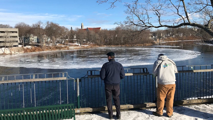 Two people stand on a river back watching a large disk of ice in a river with city buildings on the opposite bank