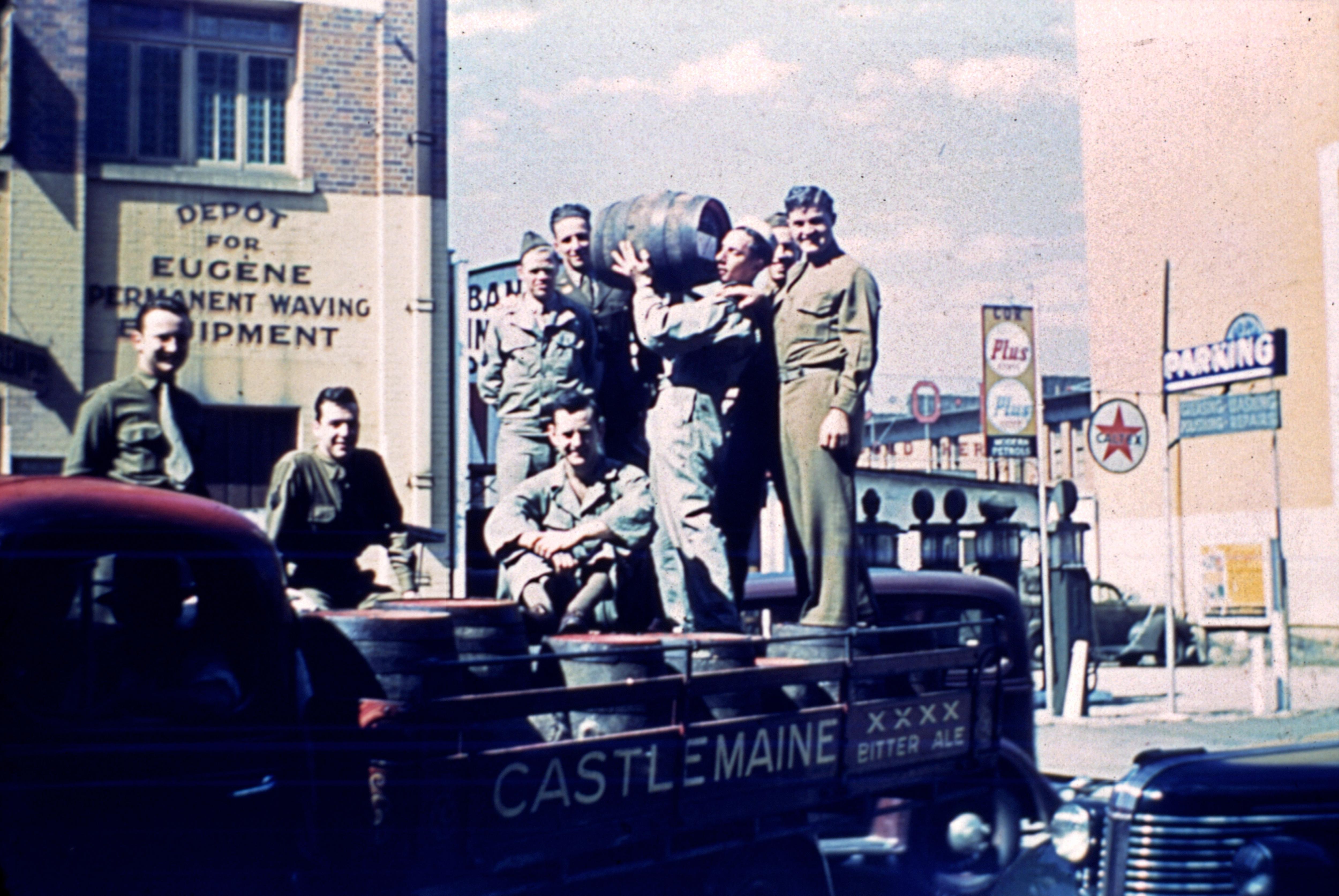 Old photo of seven US soldiers riding on the back of a truck while holding up a keg of beer.
