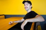 Zaynn Bird, a young man wearing a black shirt and hat, sits at a black and yellow booth in his burger shop smiling.