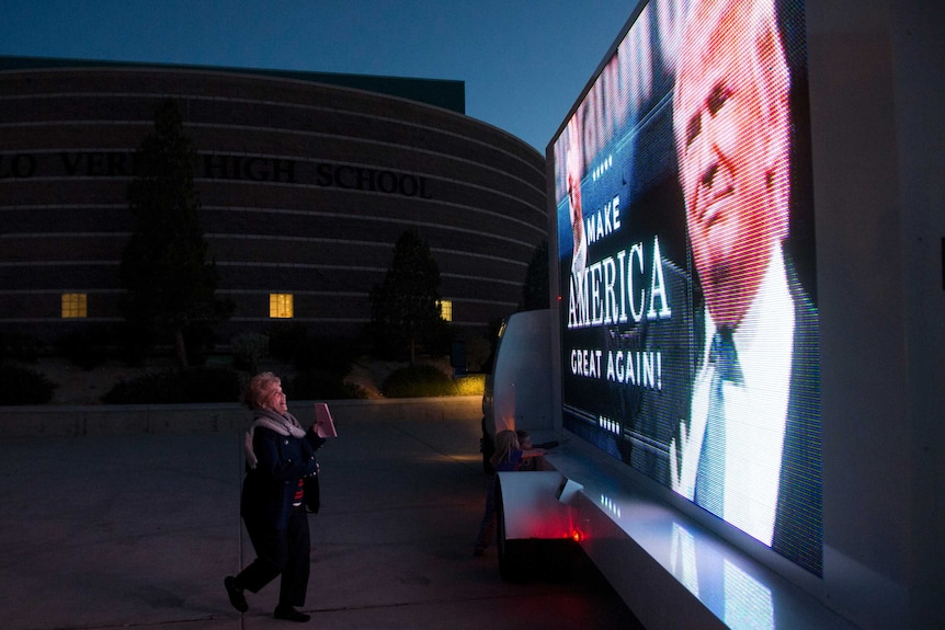 A smiling woman approaches an electronic billboard that features a picture of Donald Trump and "Make America Great Again".