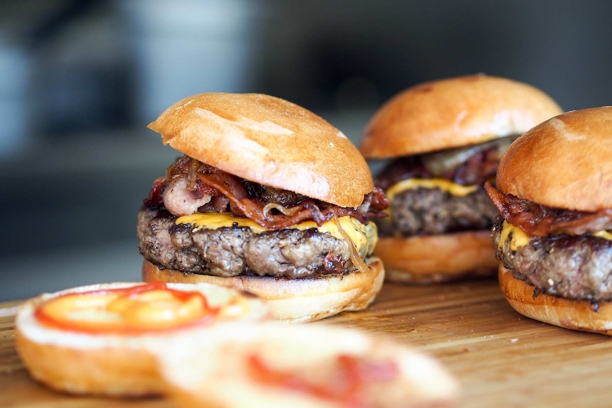 Three bacon egg cheeseburgers rest on chopping board.