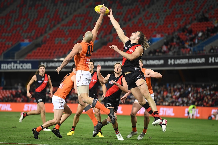 An AFL player in orange leaps into the air to contest ball possession against a player in black and red in stadium