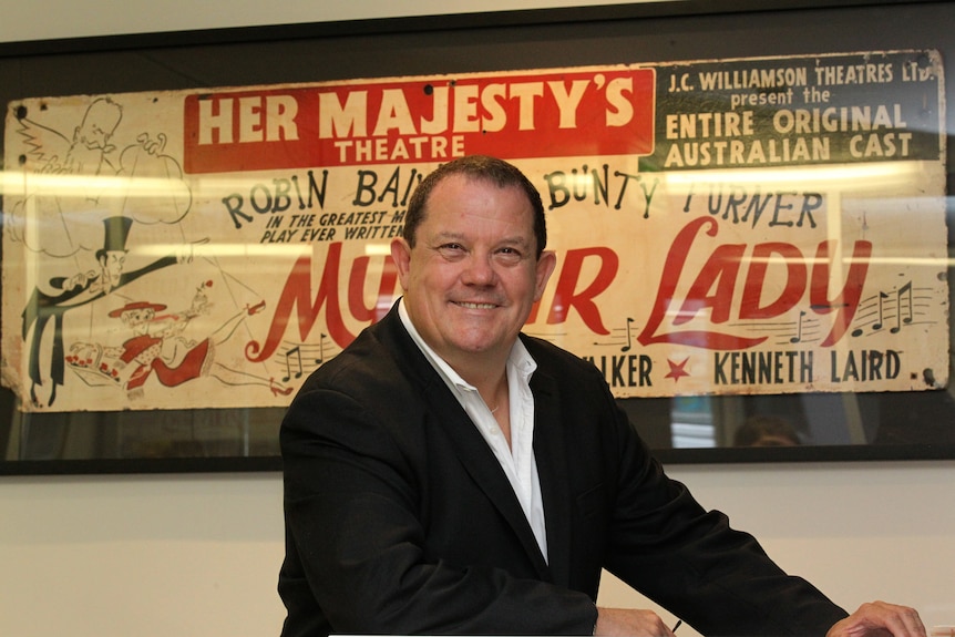A middle-aged man in a suit smiles. He is positioned in front of an ad for My Fair Lady at Her Majesty's Theatre